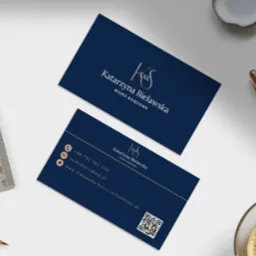 Dwie wizytówki biura księgowego Katarzyny Bielawskiej na białym biurku obok ołówka, linijki, filiżanki kawy i pojemnika z ozdobnymi kamieniami.