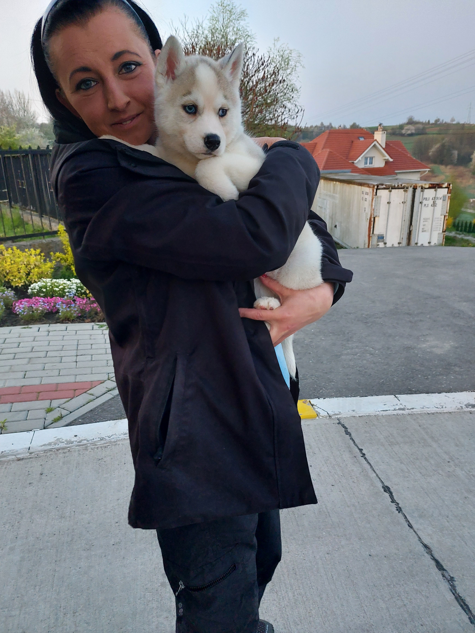 Kobieta w czarnym płaszczu trzyma na rękach szczeniaka husky o niebieskich oczach; w tle widoczny krajobraz z domem z czerwoną dachówką.