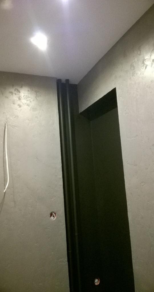 Szara ściana z efektem dekoracyjnym i czarną ościeżnicą drzwiową. Widoczne punkty instalacji elektrycznej. Oświetlenie sufitowe.