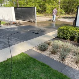 SERVICE CONSTRUCTION Sp. z o.o. - Mężczyzna w koszuli w kratę czyści ciśnieniowo kostkę brukową na podjeździe, w tle brama wjazdowa i drzewa.