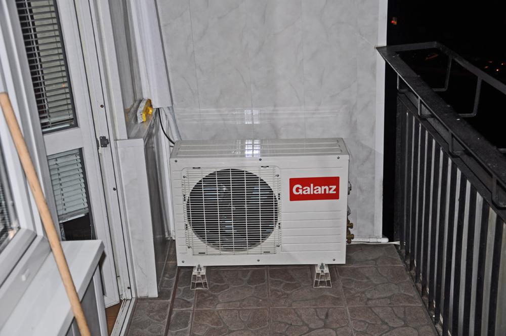 Biały agregat klimatyzacyjny Galanz zamontowany na balkonie, widoczne metalowe wsporniki i fragment balustrady.