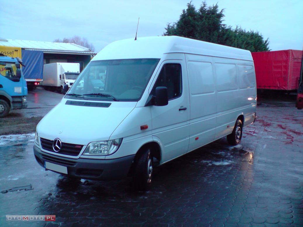 Biały, podwyższony furgon Mercedes-Benz na mokrym placu, z widocznymi innymi pojazdami transportowymi w tle.