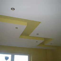 Wielopoziomowy sufit podwieszany z białych płyt z żółtymi wstawkami i punktowym oświetleniem, widok od dołu.