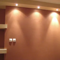 Ściana pomalowana na terakotowy kolor z trzema punktowymi lampami sufitowymi, widoczne dwa białe włączniki światła i fragment białej półki.