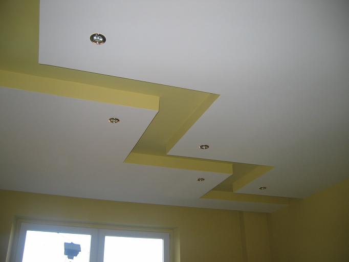 Wielopoziomowy sufit podwieszany z białych płyt z żółtymi wstawkami i punktowym oświetleniem, widok od dołu.