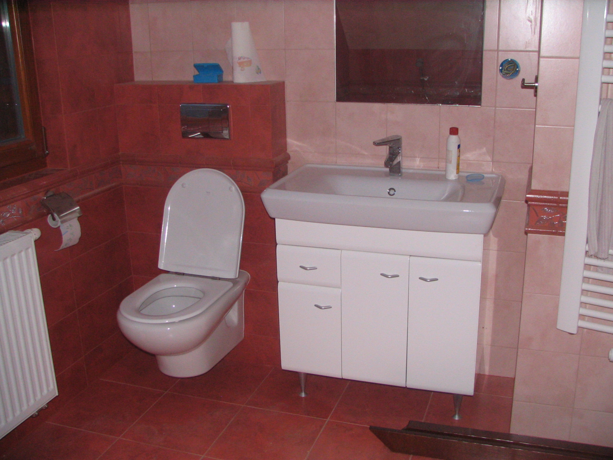 Wykończona łazienka z czerwonymi płytkami, białą toaletą i umywalką z szafką, widoczne także grzejnik i lustro.