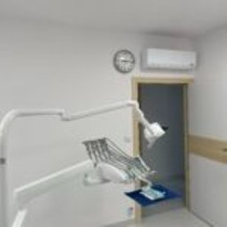 KLIMA TECHNIK, MACIEJ GLAZA - Biała klimatyzacja ścienna w gabinecie dentystycznym z zegarem i sprzętem medycznym. Widok na otwarte drzwi, oświetlenie górne.