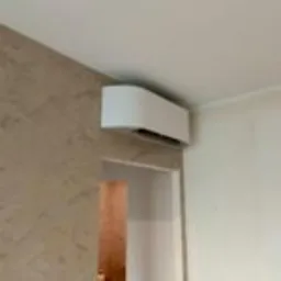 Biała klimatyzacja ścienna w jasnym wnętrzu, widok z dołu, z fragmentem drzwi po lewej stronie. Minimalistyczny design klimatyzatora.