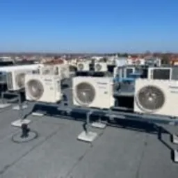 Rząd jednostek zewnętrznych klimatyzacji Panasonic na dachu budynku, zamontowanych na szarej konstrukcji wsporczej, z widokiem na miasto w słoneczny dzień.