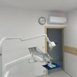 Biała klimatyzacja ścienna w gabinecie dentystycznym z zegarem i sprzętem medycznym. Widok na otwarte drzwi, oświetlenie górne.