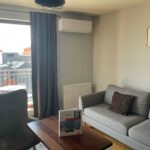 Biała klimatyzacja ścienna w jasnym salonie z szarą sofą, ciemnymi zasłonami i drewnianym stolikiem. Widok na miasto przez okno.