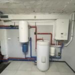 Kotłownia z dwoma białymi zbiornikami, niebieskim naczyniem przeponowym i piecem gazowym na ścianie. Rury w kolorze czerwonym i niebieskim. Wentylacja pod sufitem.