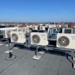 Rząd jednostek zewnętrznych klimatyzacji Panasonic na dachu budynku, zamontowanych na szarej konstrukcji wsporczej, z widokiem na miasto w słoneczny dzień.
