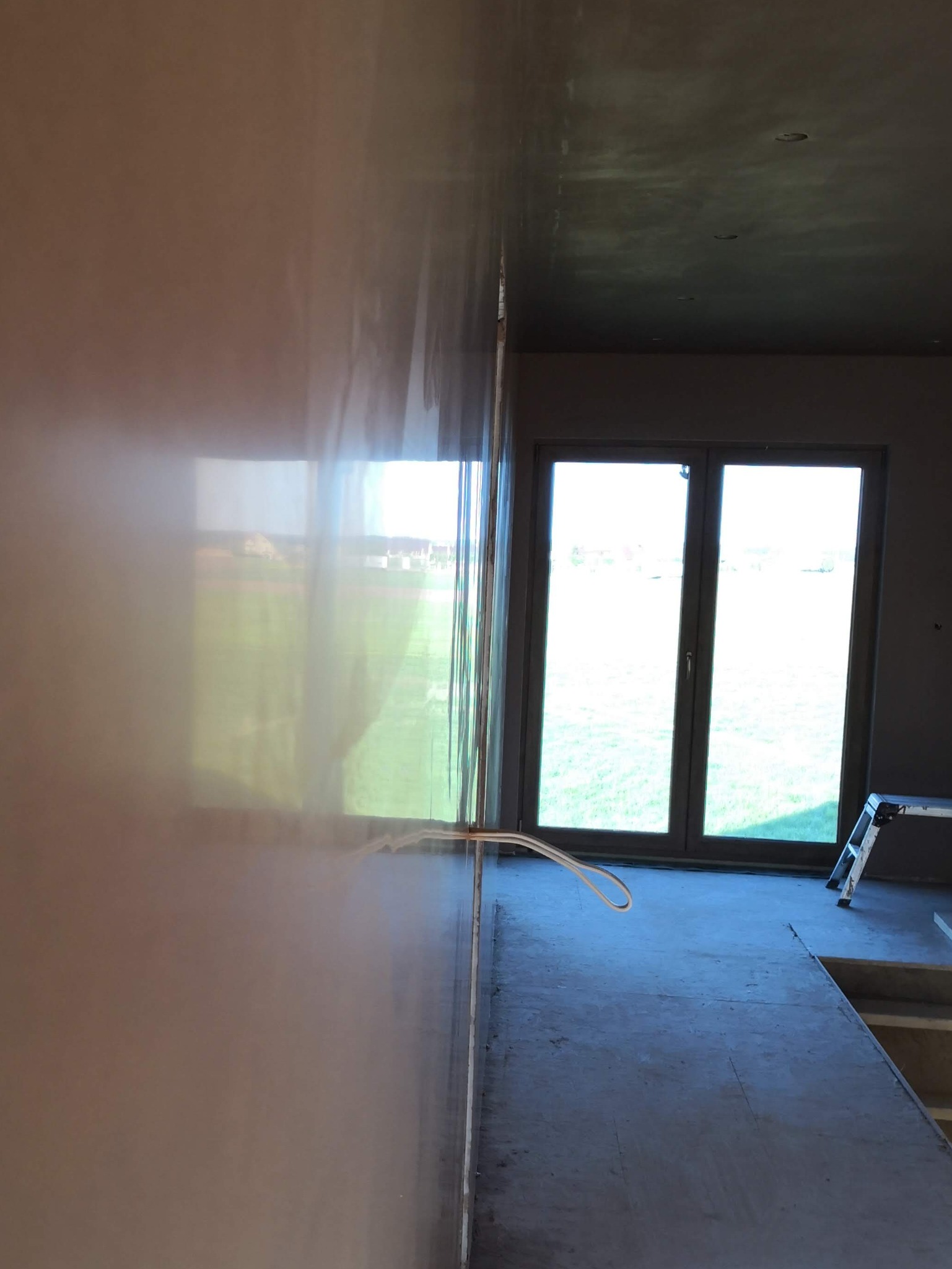 Gładka, jasna ściana po szpachlowaniu, z widocznymi przewodami elektrycznymi. W tle okno z widokiem na zieleń i drabina. Ściana w trakcie wykańczania.