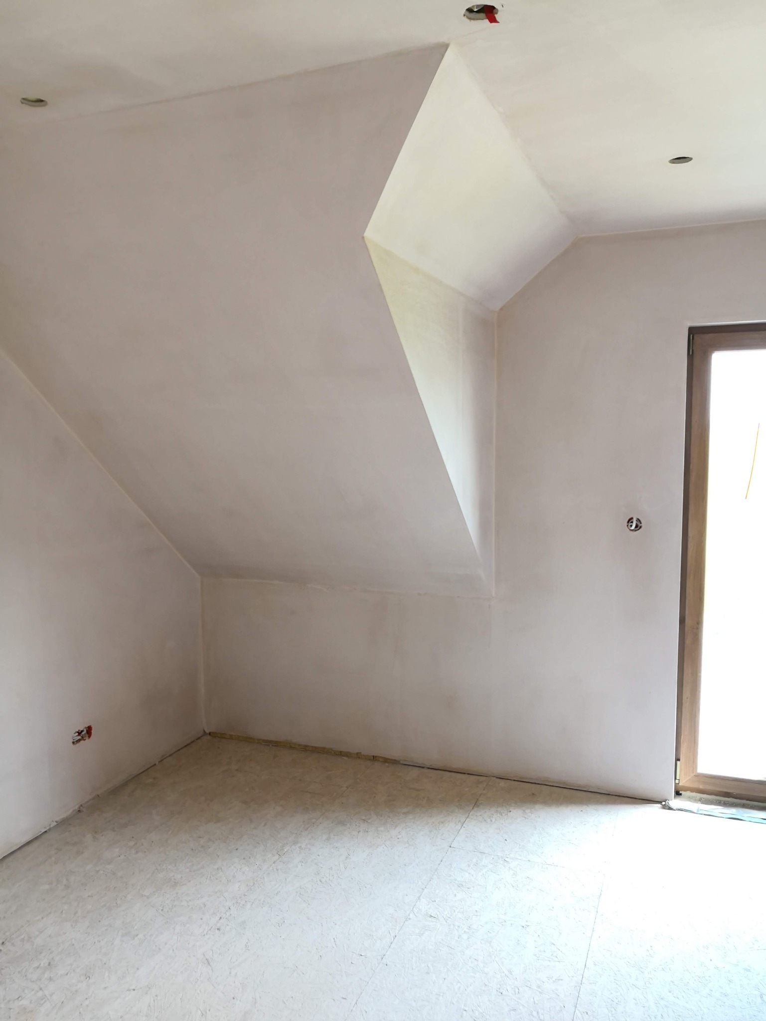 Wnętrze pokoju na poddaszu po szpachlowaniu ścian i sufitu, widoczne gładkie powierzchnie, przygotowane do malowania. Otwór okienny z widokiem na zewnątrz.