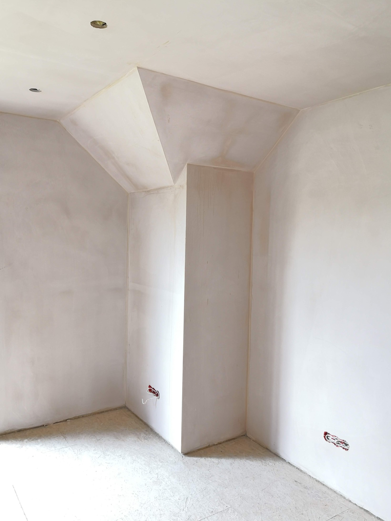 Wnętrze po szpachlowaniu ścian i sufitu, widoczne gniazdka elektryczne. Ściany w jasnym odcieniu, sufit z nietypową konstrukcją. Podłoga przygotowana do dalszych prac.