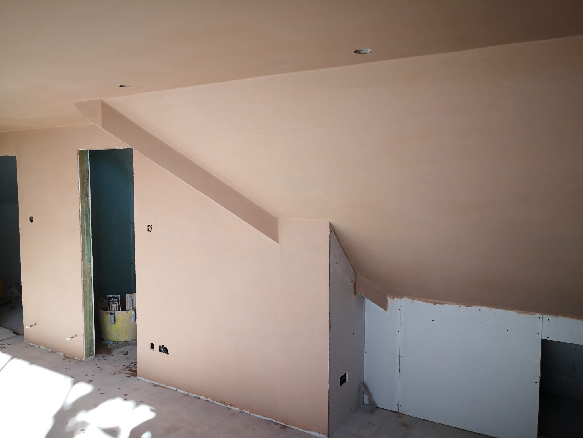 Wnętrze poddasza po szpachlowaniu ścian i sufitu na gładko, widoczne otwory na gniazdka elektryczne i fragment konstrukcji z płyt kartonowo-gipsowych.