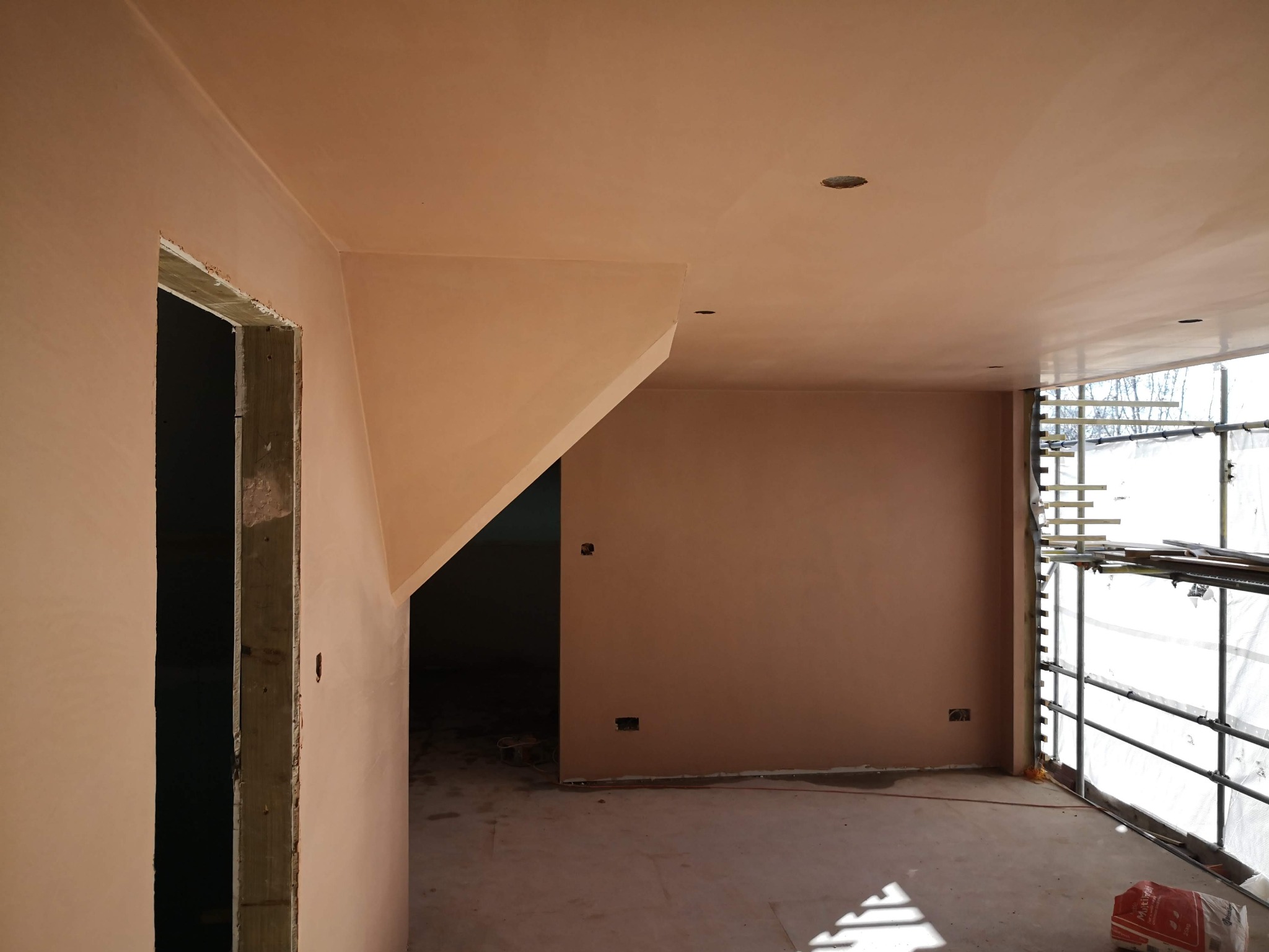 Wnętrze po szpachlowaniu: gładkie, beżowe ściany i sufit, widoczne rusztowanie przy oknie, surowa ościeżnica drzwiowa. Podłoga betonowa, przygotowana do dalszych prac.