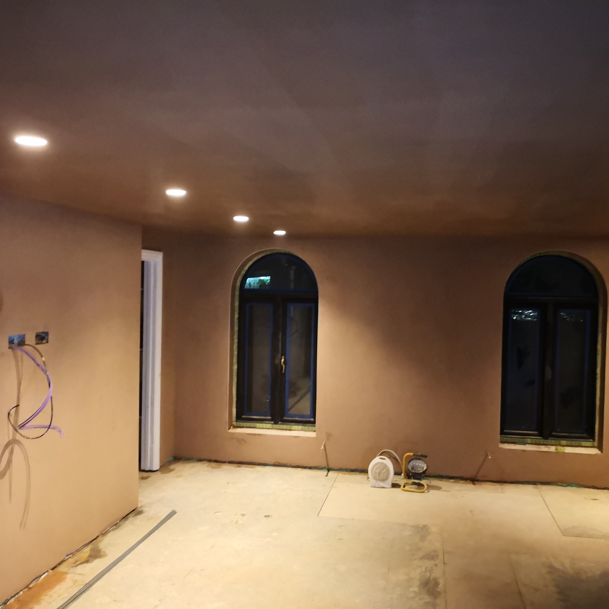 Wnętrze po szpachlowaniu: gładkie, beżowe ściany z zaokrąglonymi oknami. Widoczne kable elektryczne i lampy budowlane. Podłoga przygotowana do dalszych prac.