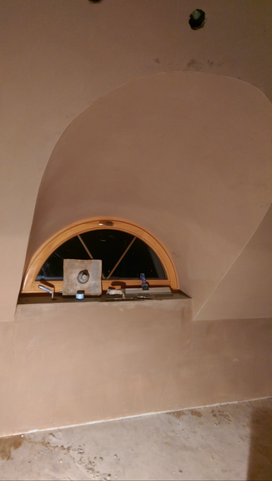 Wnętrze po szpachlowaniu: gładkie, beżowe ściany i sufit z łukowym oknem. Widoczne narzędzia szpachlarskie na parapecie. Świeżo wykończone powierzchnie.
