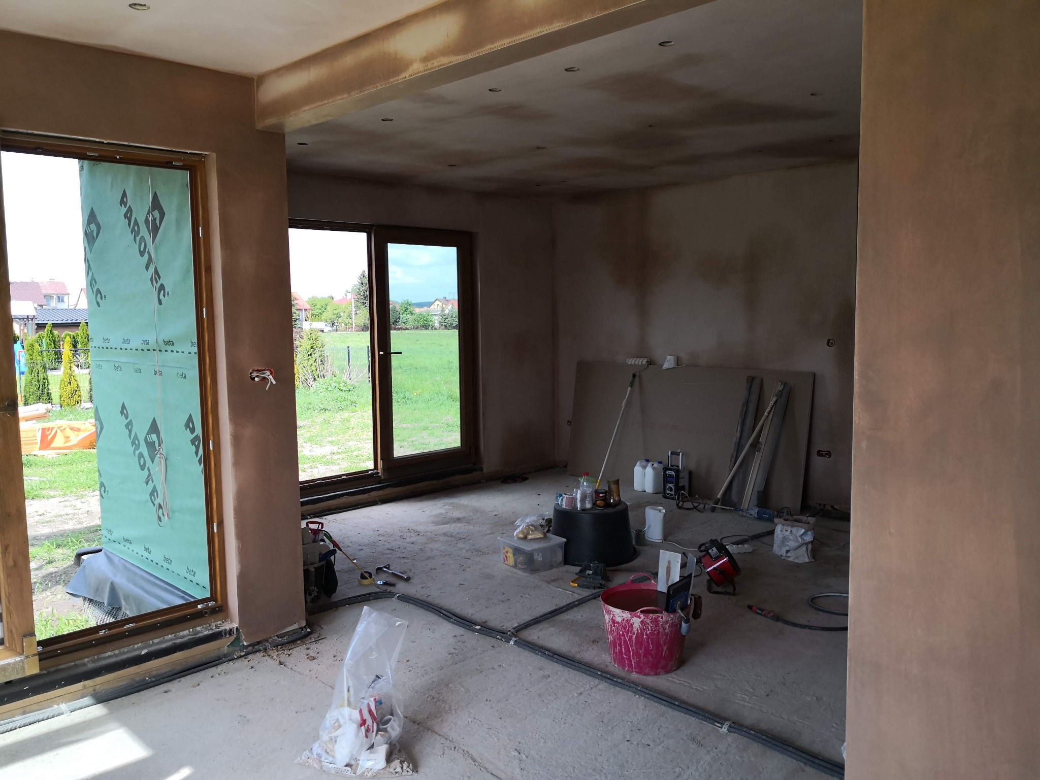 Wnętrze w trakcie prac wykończeniowych: ściany pokryte szpachlą, okna z folią ochronną Parotec, narzędzia i materiały budowlane rozłożone na podłodze. Widok na zewnątrz przez otwarte okno.
