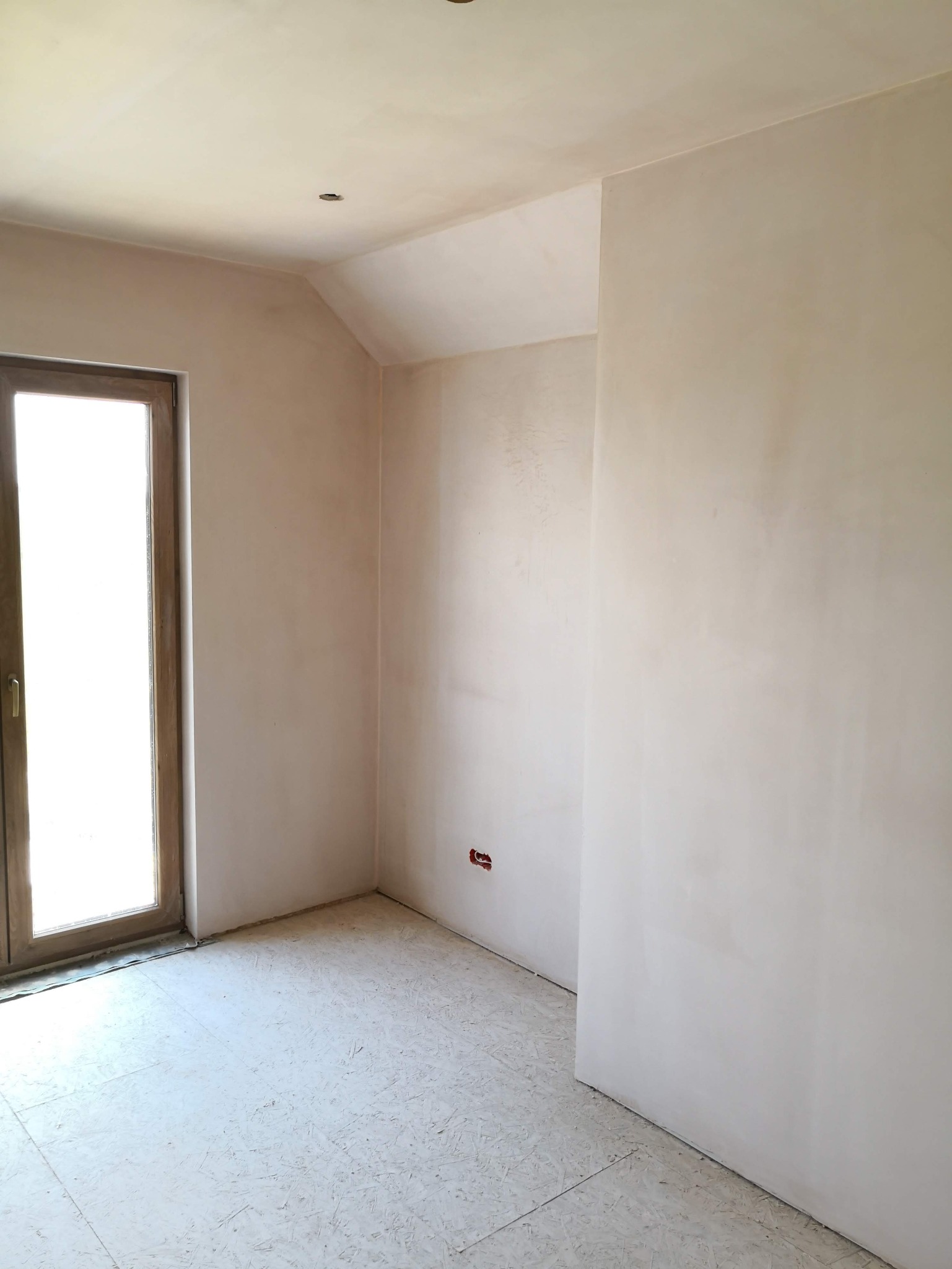 Wnętrze pokoju po szpachlowaniu ścian i sufitu, widoczne gładkie powierzchnie, okno z drewnianą ramą i przygotowane podłoże. Świeżo wykończone wnętrze.