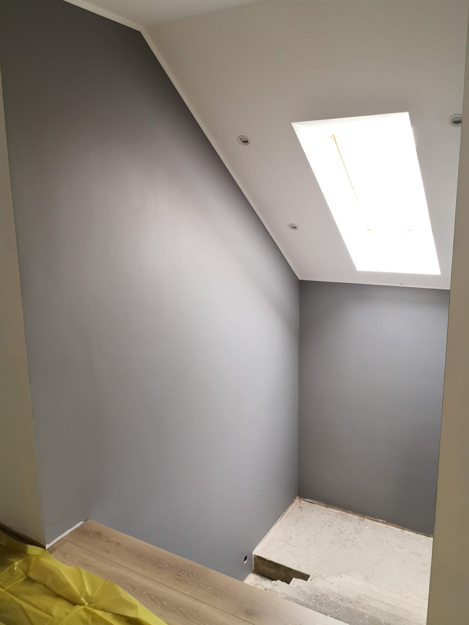 Wnętrze po malowaniu ścian na szaro, widoczne schody z zabezpieczoną podłogą i sufit z oknem dachowym. Minimalistyczny design.