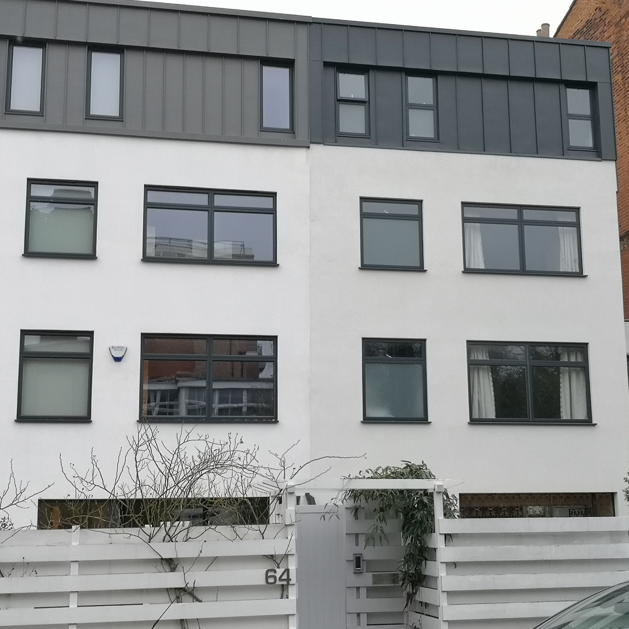 Biała fasada bliźniaka z szarym dachem i ciemnymi oknami. Widoczny fragment białego płotu z numerem 64 i roślinnością. Minimalistyczny design.