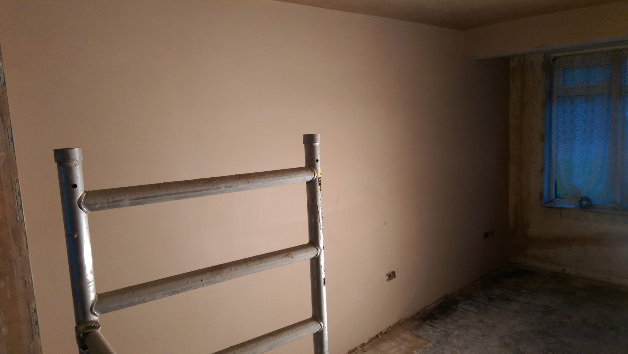 Wnętrze po szpachlowaniu ścian, widoczne rusztowanie malarskie i okno z zasłoną. Ściany gładkie, przygotowane do malowania. Podłoga w trakcie prac wykończeniowych.