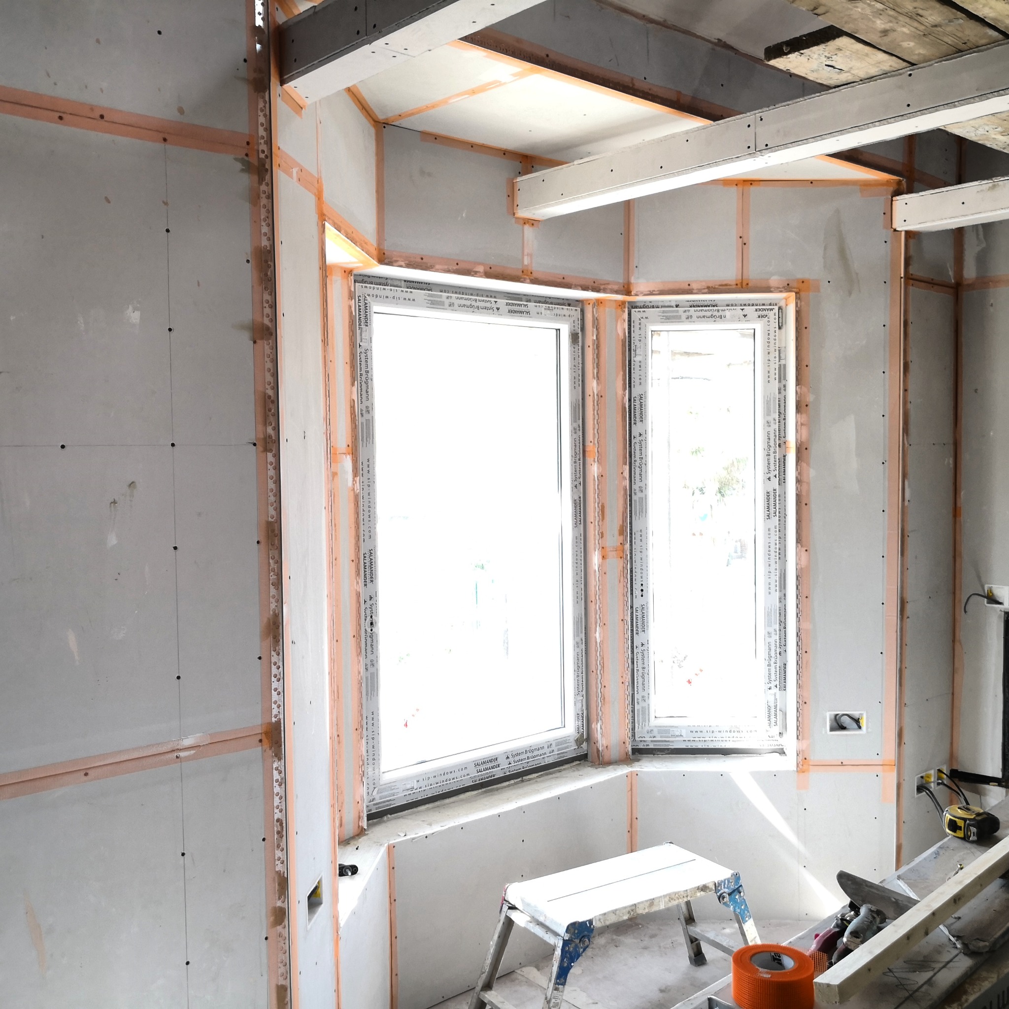 Wnętrze w trakcie wykańczania: płyty gipsowo-kartonowe na ścianach, okna z widocznymi oznaczeniami producenta, taśma wzmacniająca na łączeniach, drabina i narzędzia.