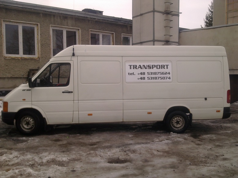 Biały samochód dostawczy z napisem TRANSPORT i numerami telefonów na boku, zaparkowany na błotnistym terenie z resztkami śniegu, na tle szarego budynku.