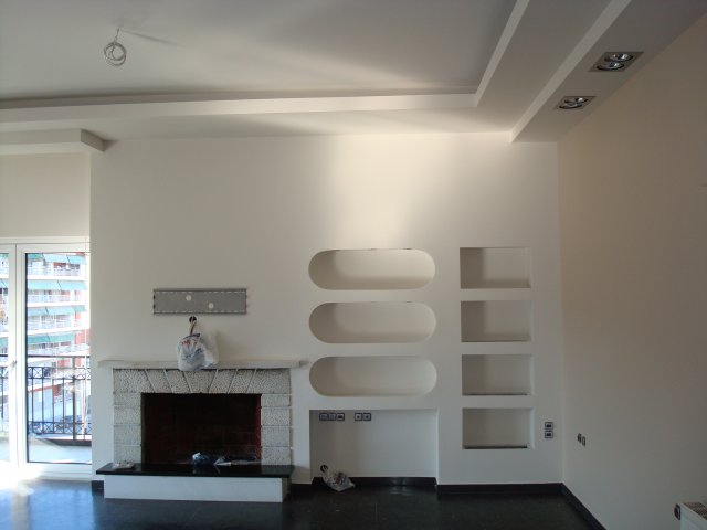 Wnętrze pokoju z kominkiem o obudowie z jasnych kamieni i wnękami w ścianie, widoczne przygotowania do montażu sufitu podwieszanego z płyt gipsowo-kartonowych, częściowo oświetlone światłem...