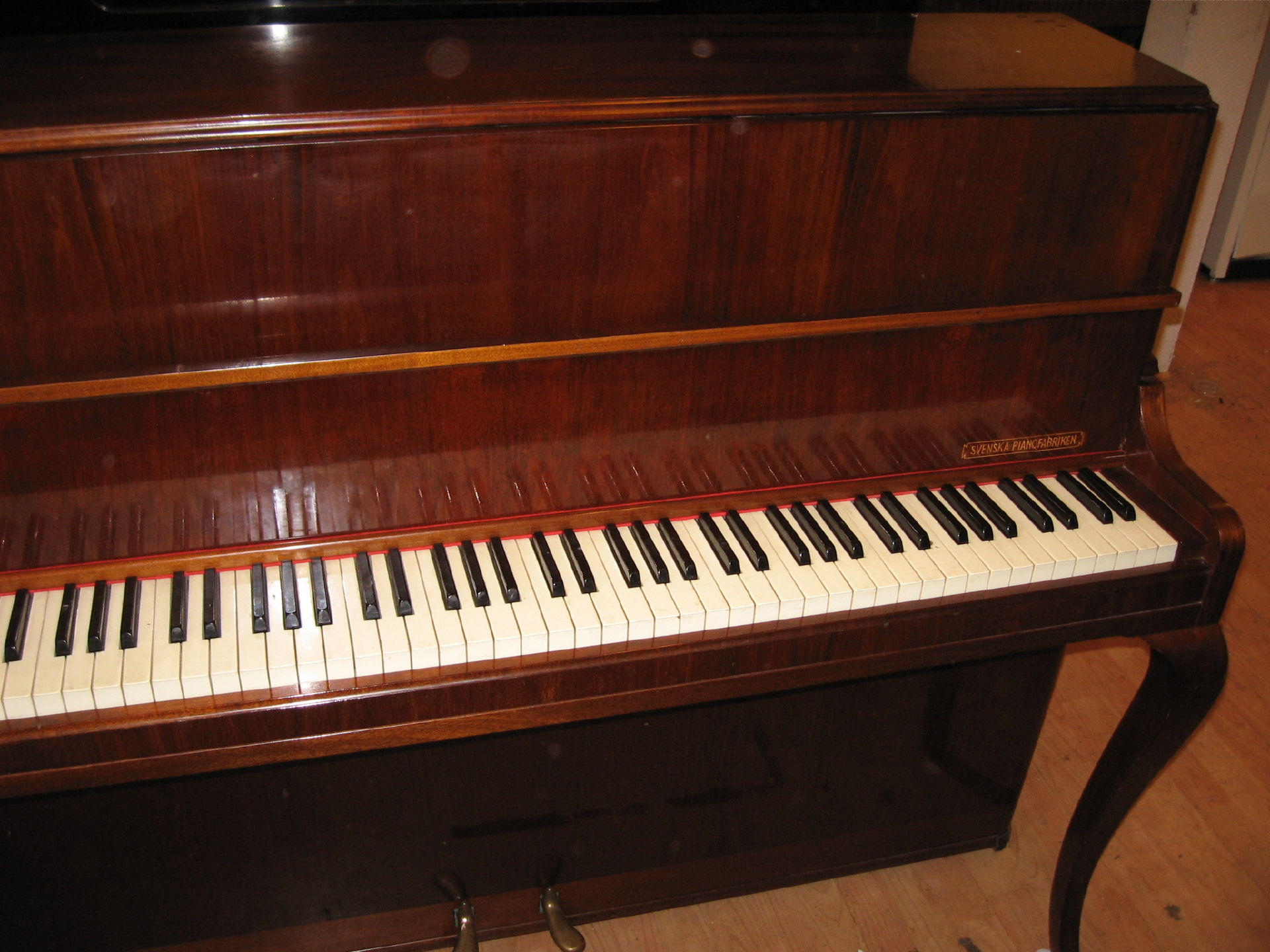 Pionowy fortepian o mahoniowym wykończeniu z widocznym napisem 'SVENSKA PIANOFABRIKEN' nad klawiaturą.