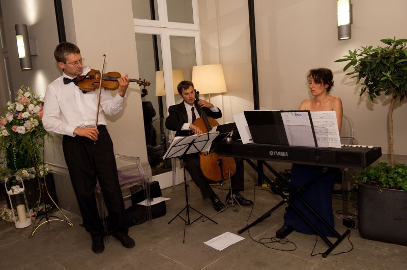 Zespół muzyczny: skrzypek w muszce, wiolonczelista w garniturze i pianistka grająca na keyboardzie Yamaha podczas uroczystości.