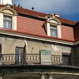 Fragment elewacji budynku z balkonem i dachem pokrytym czerwoną dachówką karpiówką, widoczne okna dachowe z ozdobnymi obramowaniami.