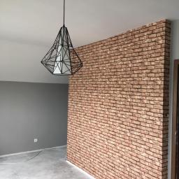 Veldecor - Ściana wykończona tapetą imitującą czerwoną cegłę, oświetlona nowoczesną lampą wiszącą z geometrycznym kloszem, na tle szarej ściany i surowej podłogi.