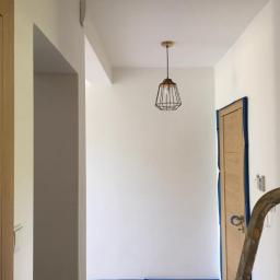 Veldecor - Świeżo pomalowany, jasny korytarz z drewnianymi drzwiami zabezpieczonymi taśmą malarską, widoczna metalowa lampa wisząca i fragment drabiny malarskiej.
