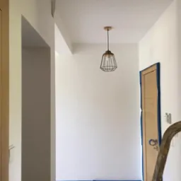 Świeżo pomalowany, jasny korytarz z drewnianymi drzwiami zabezpieczonymi taśmą malarską, widoczna metalowa lampa wisząca i fragment drabiny malarskiej.