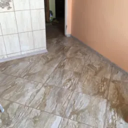 Ułożone, beżowo-brązowe płytki podłogowe imitujące marmur w rogu pokoju z pomarańczową ścianą i otwartymi drzwiami do innego pomieszczenia.