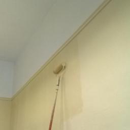 Veldecor - Świeżo pomalowana ściana w odcieniach beżu z widoczną linią odcięcia koloru przy suficie, z wałkiem malarskim i fragmentem rusztowania w kadrze.