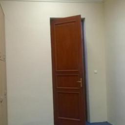 Veldecor - Pokój z beżowymi ścianami, niebieską wykładziną i częściowo otwartymi, brązowymi drzwiami z trzema panelami, oświetlony górnym światłem.