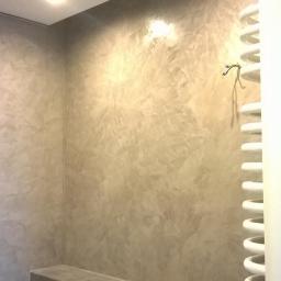 Veldecor - Ściana w łazience wykończona dekoracyjną, szarą gładzią w stylu weneckim, z widocznym grzejnikiem drabinkowym i fragmentem szafki.
