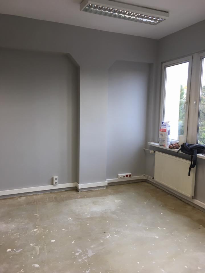 Pokój po świeżym pomalowaniu na szaro, widoczne ślady farby na podłodze, grzejnik pod oknem, worek z materiałem budowlanym na parapecie.