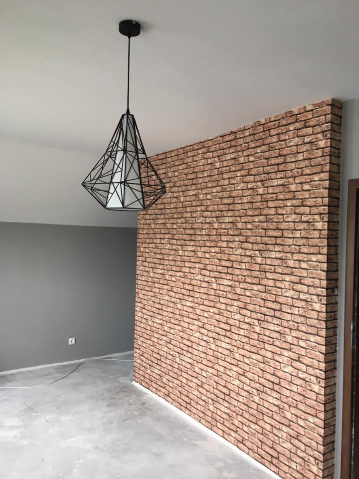 Ściana wykończona tapetą imitującą czerwoną cegłę, oświetlona nowoczesną lampą wiszącą z geometrycznym kloszem, na tle szarej ściany i surowej podłogi.