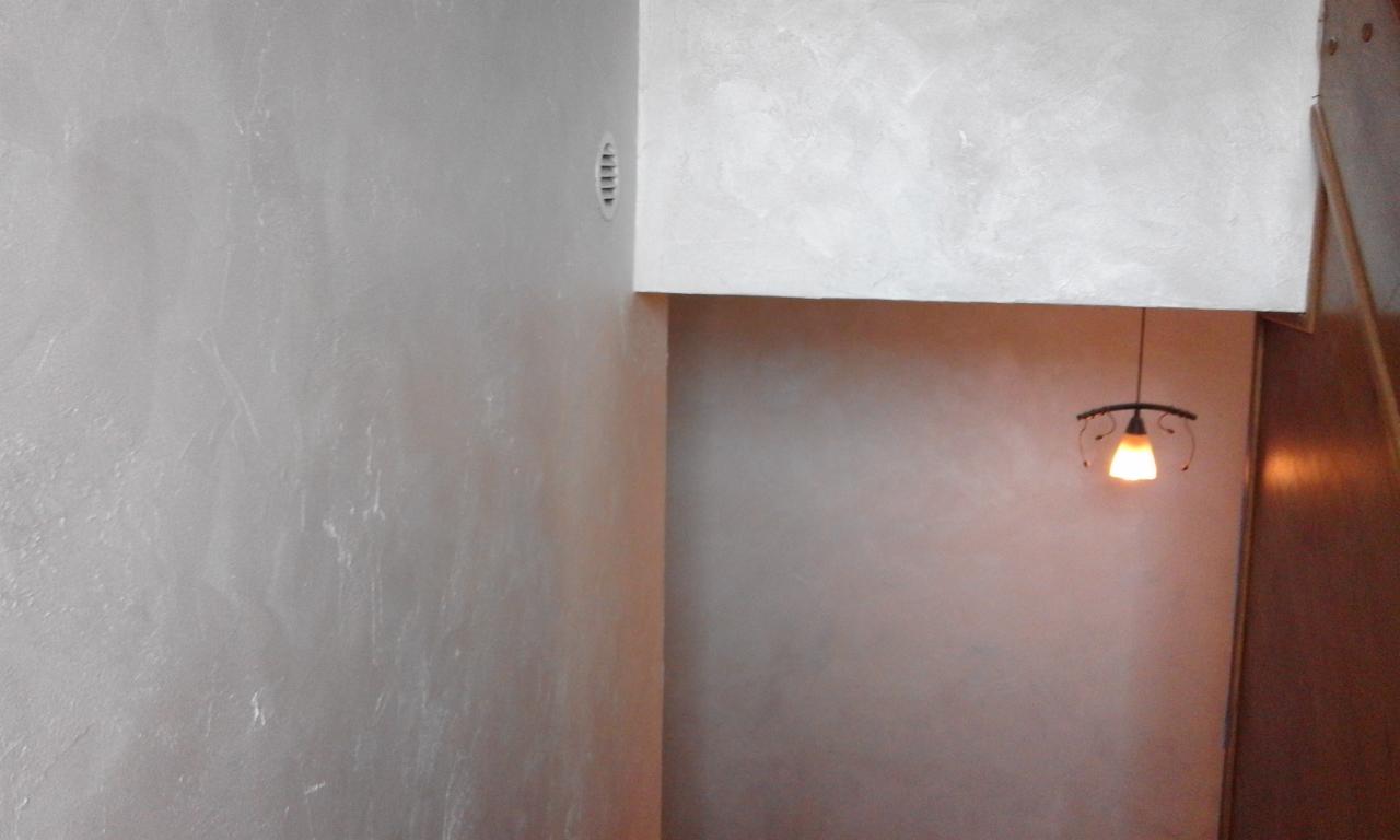 Ściana pokryta dekoracyjną, srebrzystą powłoką z widoczną fakturą, oświetlona ciepłym światłem lampy wiszącej, z kratką wentylacyjną w rogu.