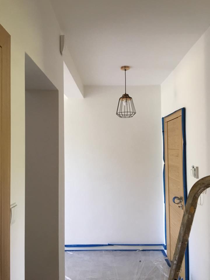 Świeżo pomalowany, jasny korytarz z drewnianymi drzwiami zabezpieczonymi taśmą malarską, widoczna metalowa lampa wisząca i fragment drabiny malarskiej.