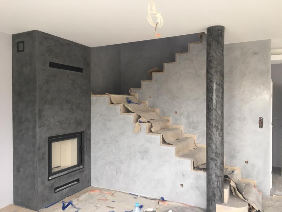 Ściana z kominkiem i schody w trakcie wykańczania gładzią dekoracyjną w odcieniach szarości, zabezpieczone schody i podłoga.