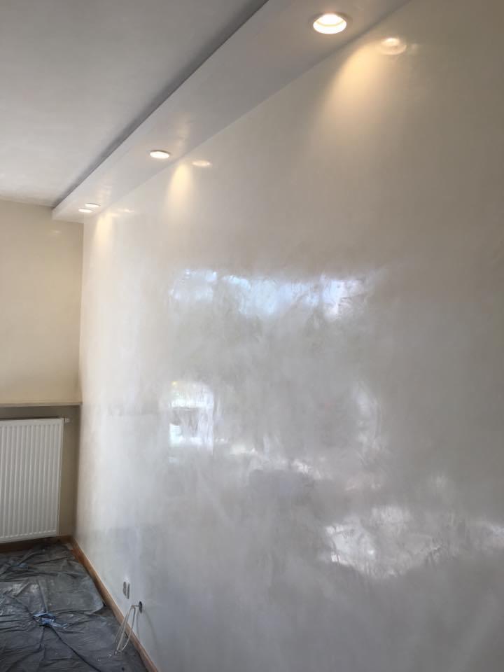 Ściana wykończona gładzią w odcieniach bieli i szarości, oświetlona punktowymi lampami sufitowymi, z widocznym grzejnikiem i zabezpieczoną folią podłogą.