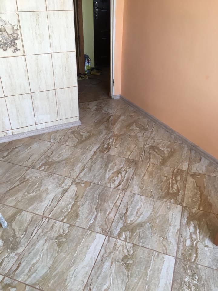 Ułożone, beżowo-brązowe płytki podłogowe imitujące marmur w rogu pokoju z pomarańczową ścianą i otwartymi drzwiami do innego pomieszczenia.