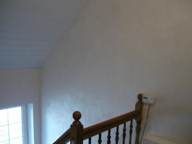 Ściana w klatce schodowej po nałożeniu gładzi, widoczna drewniana balustrada z okrągłymi zakończeniami słupków i regulator temperatury na ścianie.